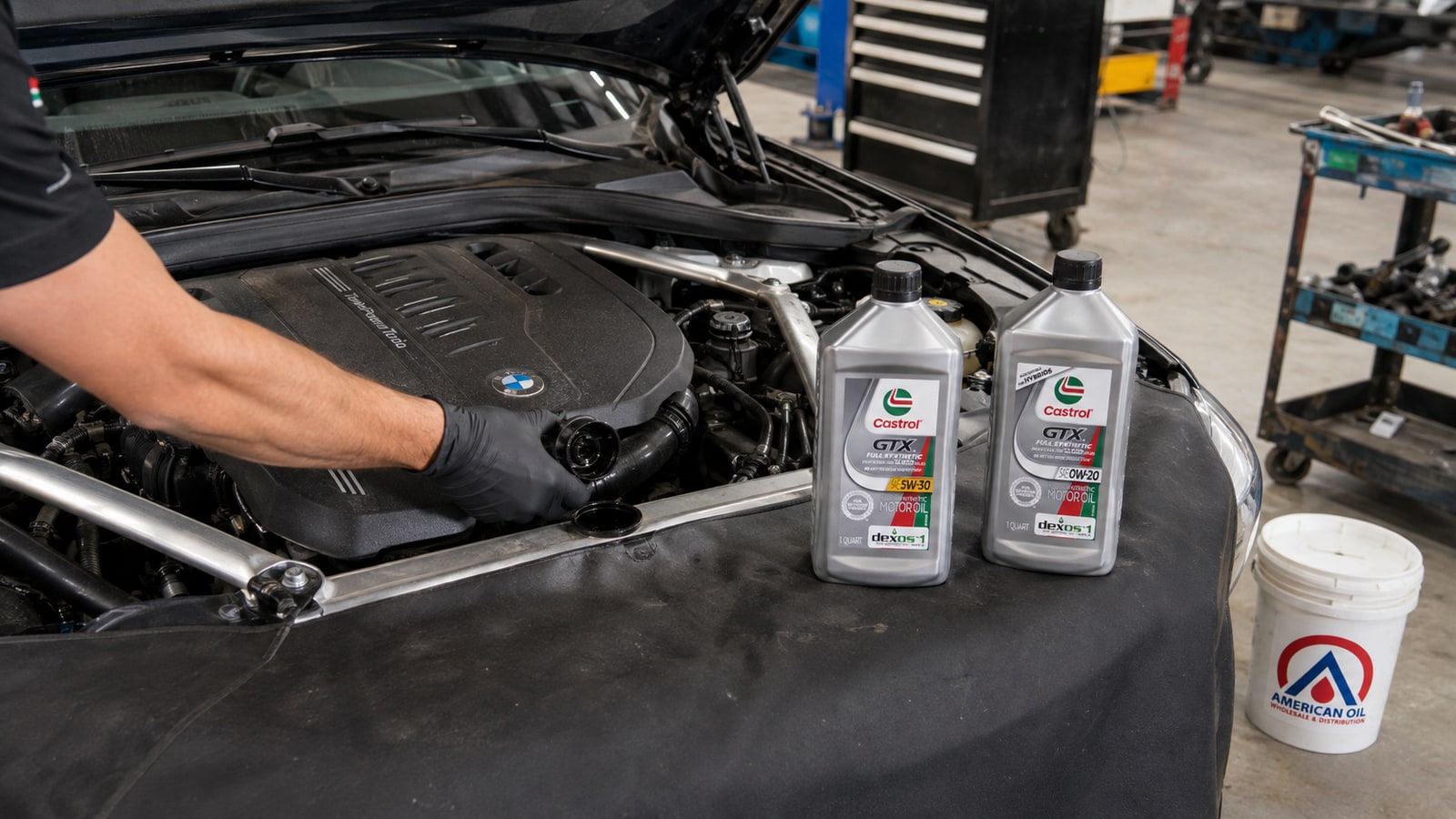 Technician opening the oil filler cap on a European car engine with Castrol full synthetic quarts and a bulk pail in a South Florida shop